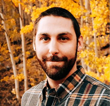 A man with a beard and short hair, wearing a plaid shirt, stands outdoors in front of trees with yellow autumn leaves.