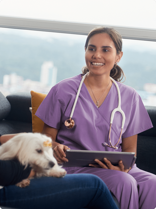 A veterinarian in purple scrubs with a stethoscope smiles while holding a tablet, sitting near a small white dog on a person's lap.