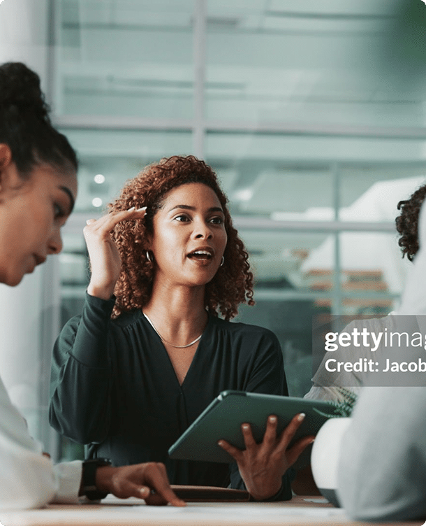 A woman holding a tablet gestures while speaking to two colleagues in a modern office meeting room.