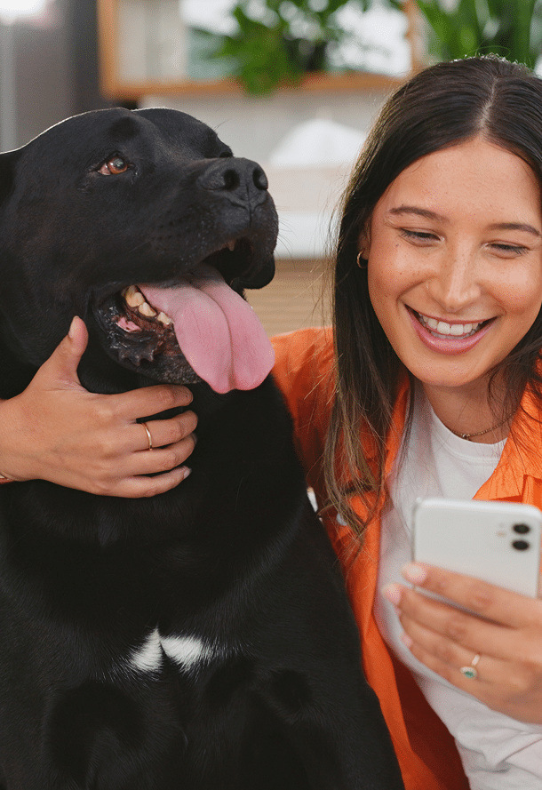 A woman in an orange shirt takes a selfie with a large black dog indoors. Both appear relaxed and happy.