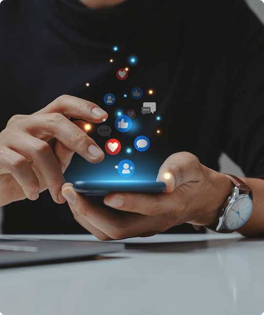 Person using a smartphone with digital social media icons, such as thumbs up, chat bubbles, and hearts, appearing above the device.