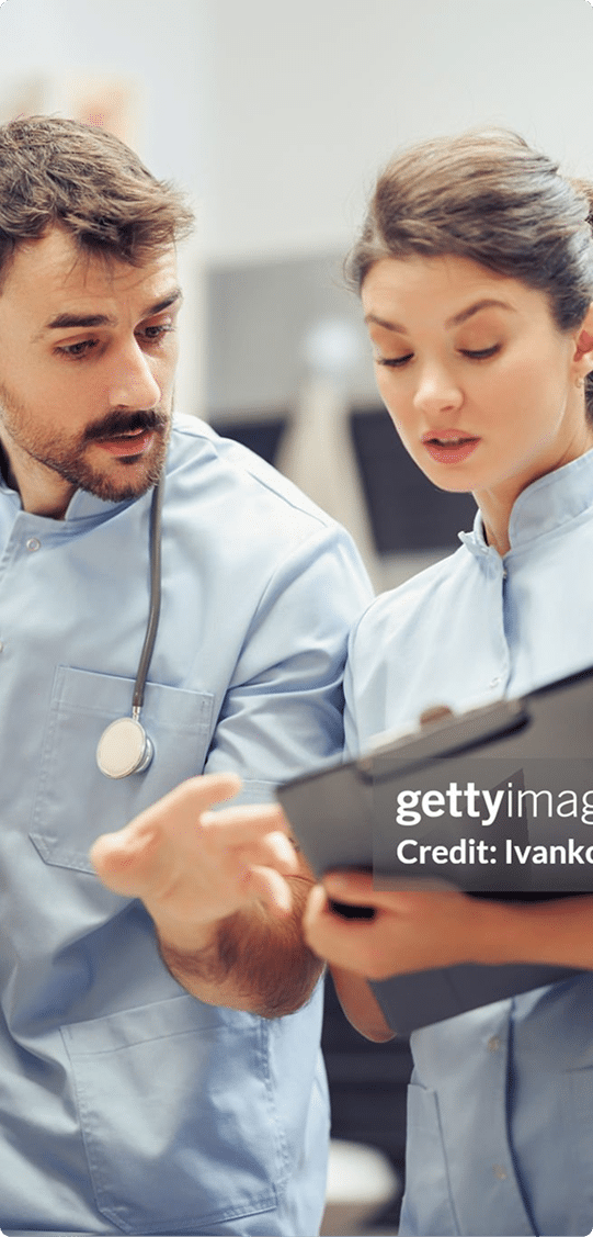 Two healthcare professionals in scrubs discuss information on a clipboard in a medical setting.