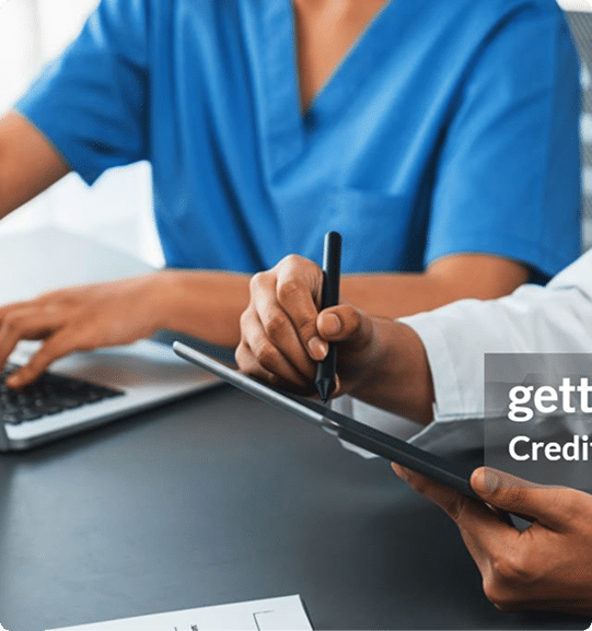 Two people are seated at a table; one wears blue scrubs and types on a laptop, while the other uses a stylus to write on a tablet.