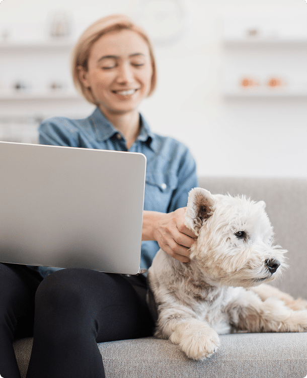 A person sitting on a couch using a laptop, while petting a small white dog lying beside them.