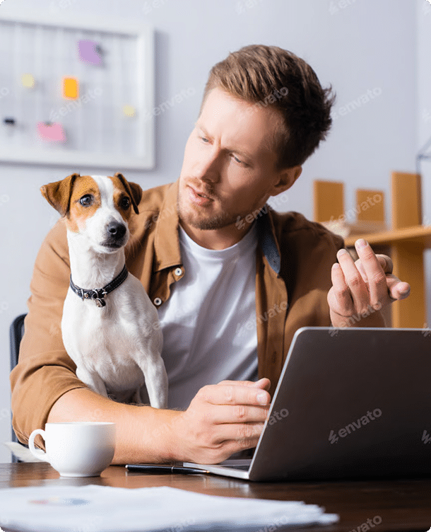 Man sitting at a desk with a laptop and coffee cup, looking at a small dog sitting beside him, in a home office setting.