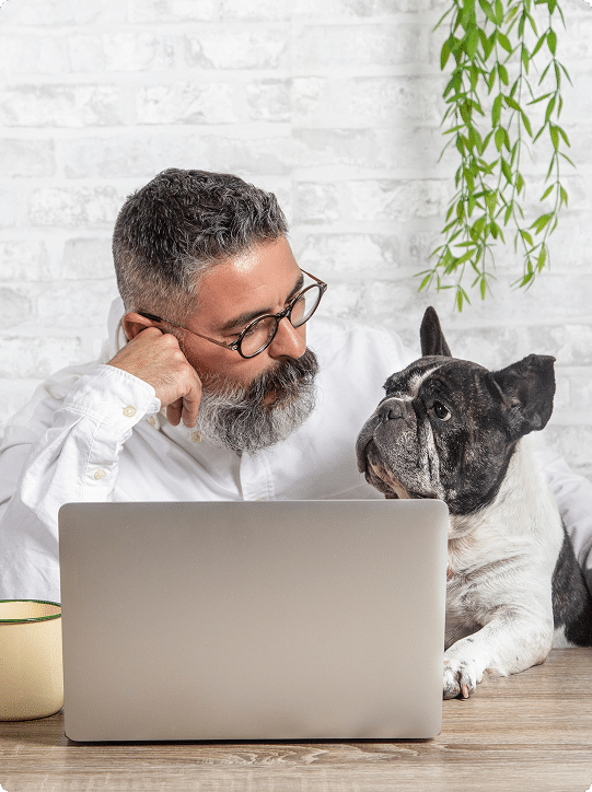 A man with glasses and a beard sits at a desk with a laptop, looking at a French Bulldog beside him. A coffee mug and hanging plant are visible in the background.